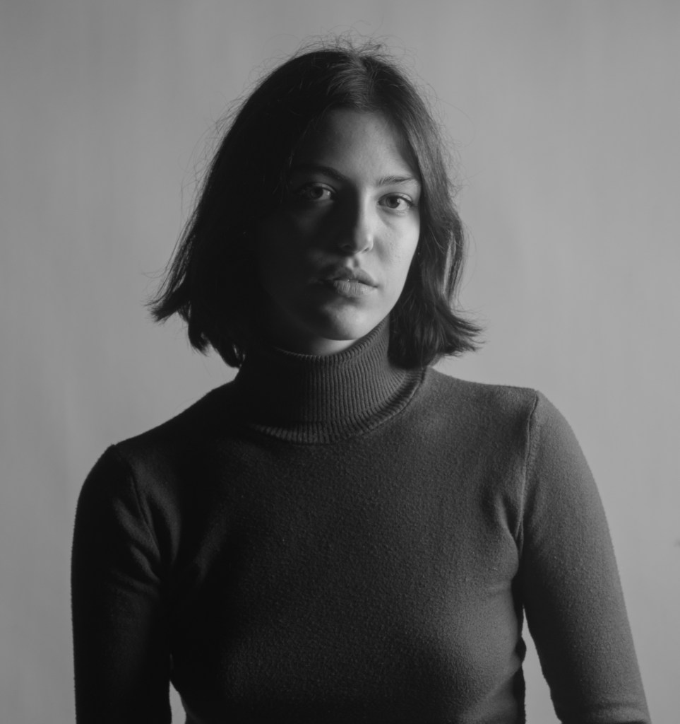 A monochrome portrait of artist Athina Alkini with shoulder-length hair wearing a turtleneck sweater, facing the camera with a neutral expression against a plain background.
